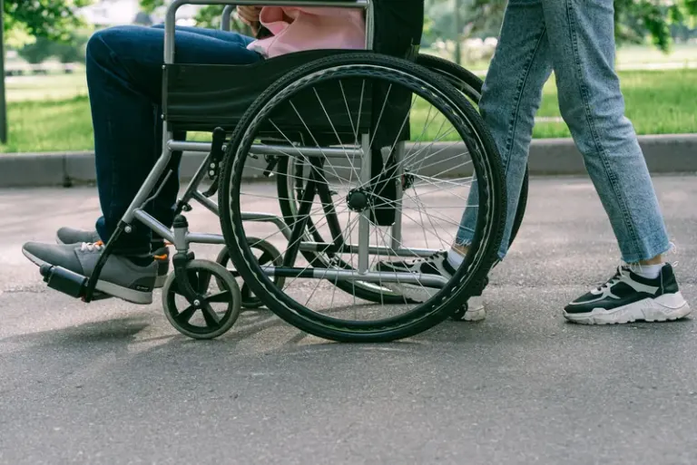 Close-up view of a person in a wheelchair being assisted on a sunny urban pavement. Stock Photo