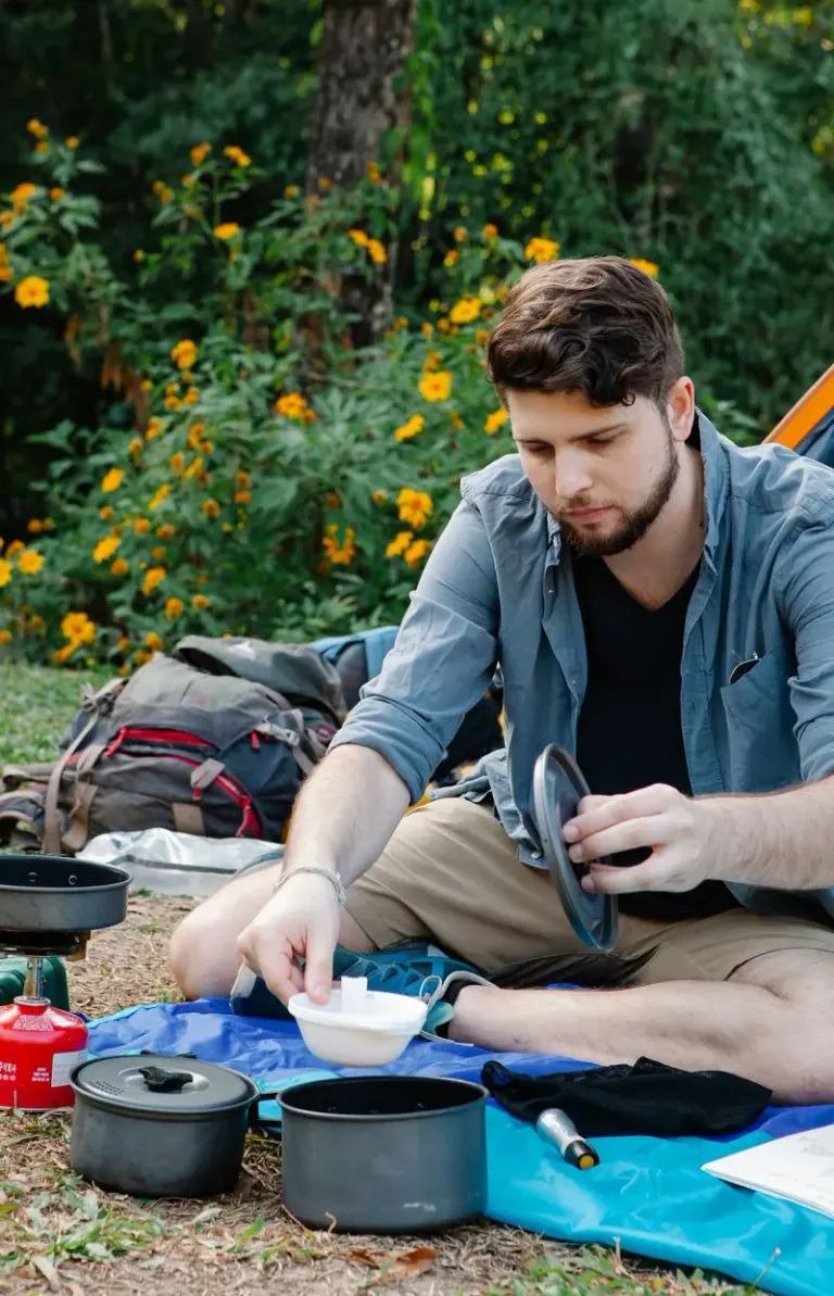 Concentrated young bearded male hiker in casual clothes pouring water into saucepan while preparing food during camping in nature Stock Photo