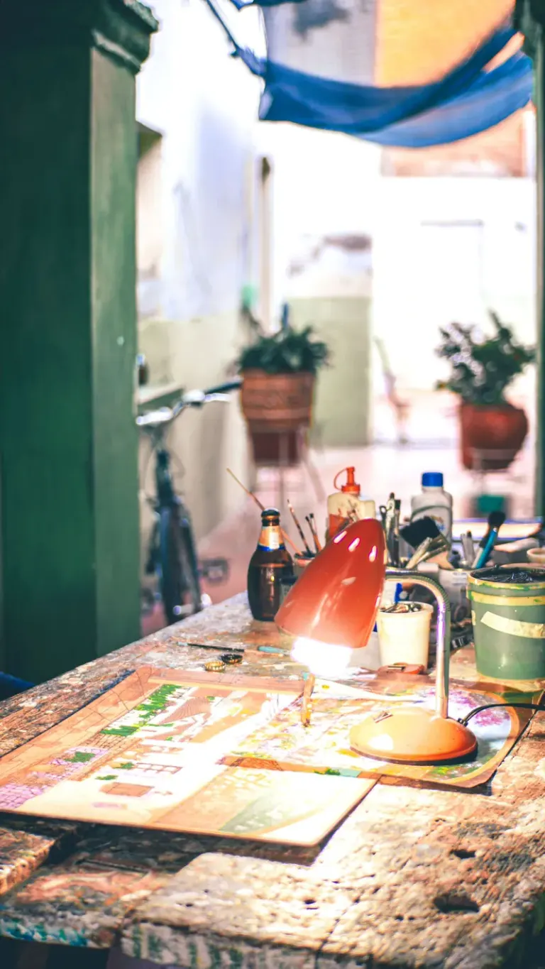 A well-lit artistic workspace featuring a rustic desk, lamp, and creative tools in an informal setting. Stock Photo