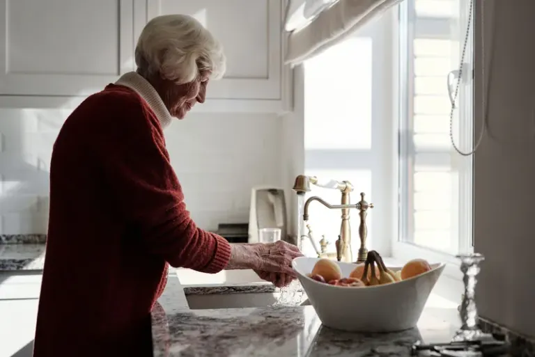 Elderly woman washing fruits at a kitchen sink bathed in sunlight. Stock Photo