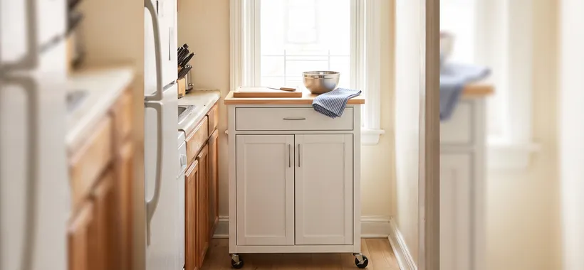 Slim rolling kitchen island cart parked on the narrow floor slice beside the counter of a small galley kitchen, walkway still clear