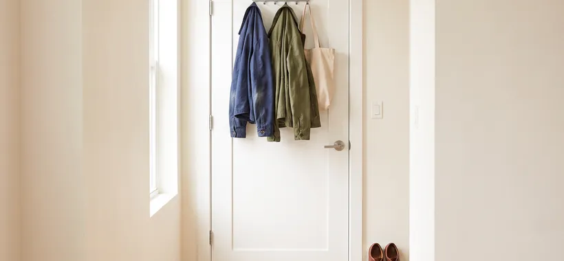 A slim hook rack hangs over the top edge of an entryway door, holding two jackets and a tote bag while the small floor below stays clear.